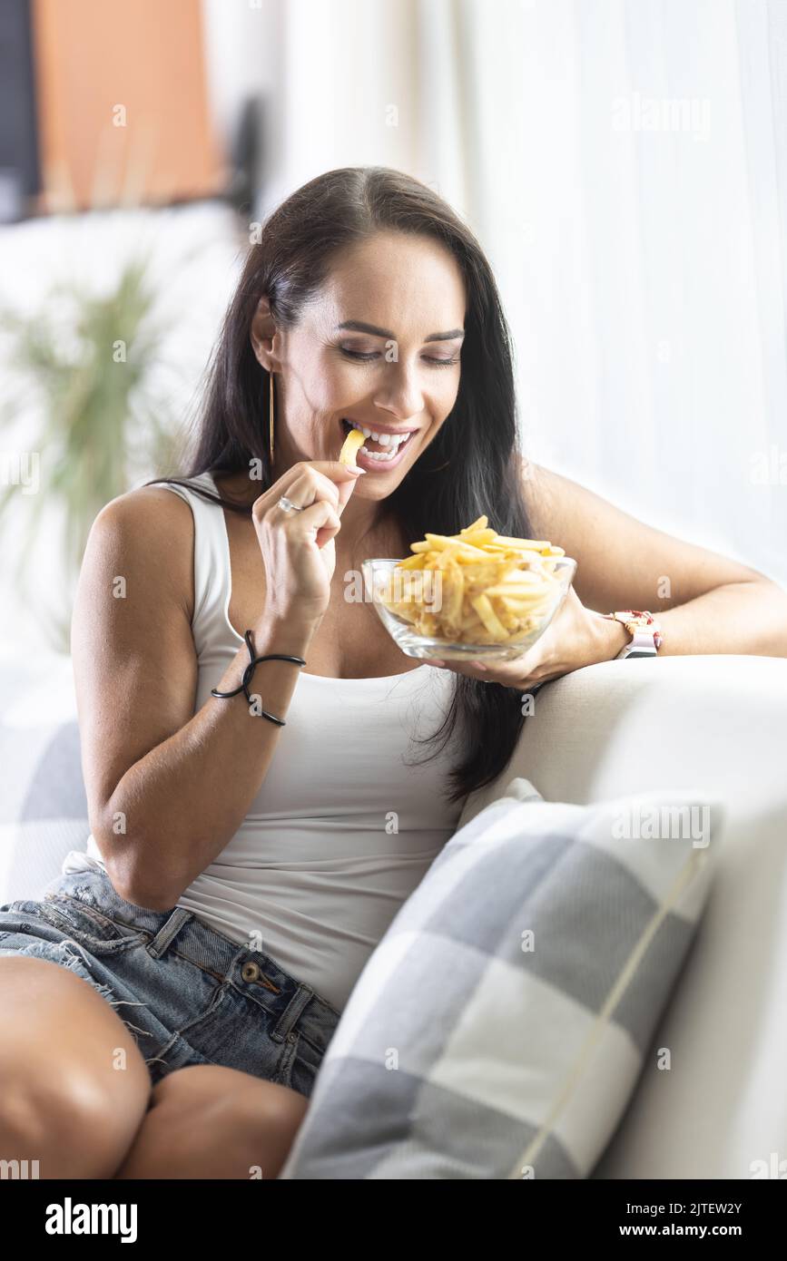 Lächelnde Frau beißt in eine der pommes, die aus einer Glasschüssel genommen werden, während sie zu Hause auf dem Sofa sitzt. Stockfoto