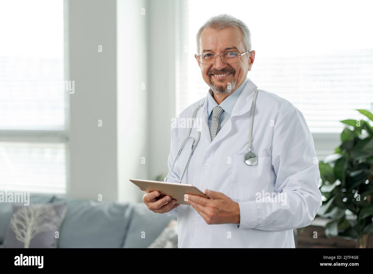 Porträt eines älteren Arztes, er schaut in die Kamera. Der Arzt steht vor dem Hintergrund einer medizinischen Klinik und hält eine Tablette in den Händen. Stockfoto