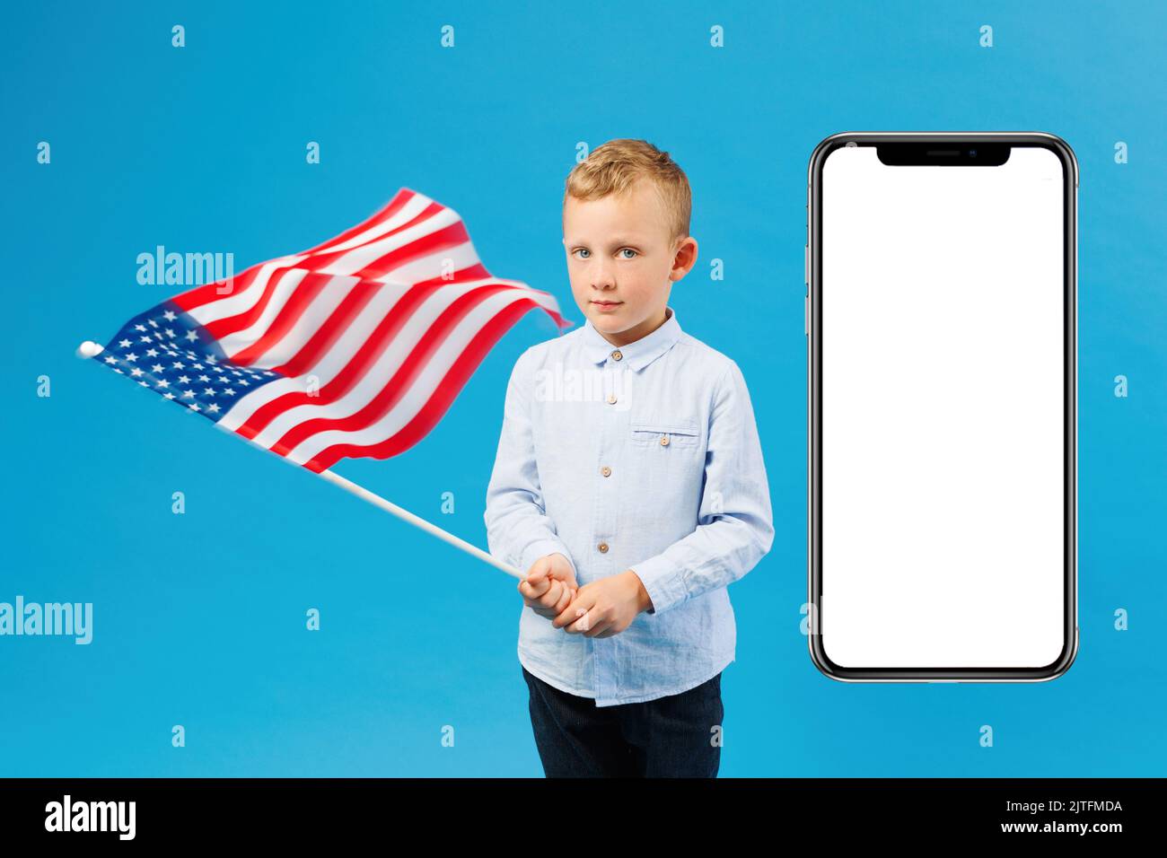 Netter Junge mit amerikanischer Flagge im Studio auf blauem Hintergrund in der Nähe eines riesigen Mobiltelefons. Das Konzept des Patriotismus gegenüber dem Land Stockfoto