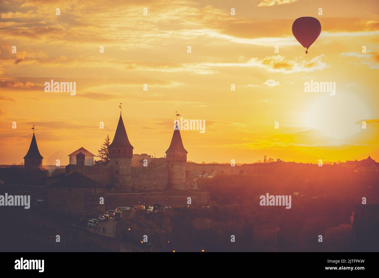 Strahlend orangefarbener Sonnenuntergang über der Burg Kamianets-Podilskyi. Ein Heißluftballon stellt sich vor dem goldenen Himmel und beobachtet das Stadtbild der Altstadt am Abend. Reisehintergrund. Beliebtes Touristenziel Stockfoto