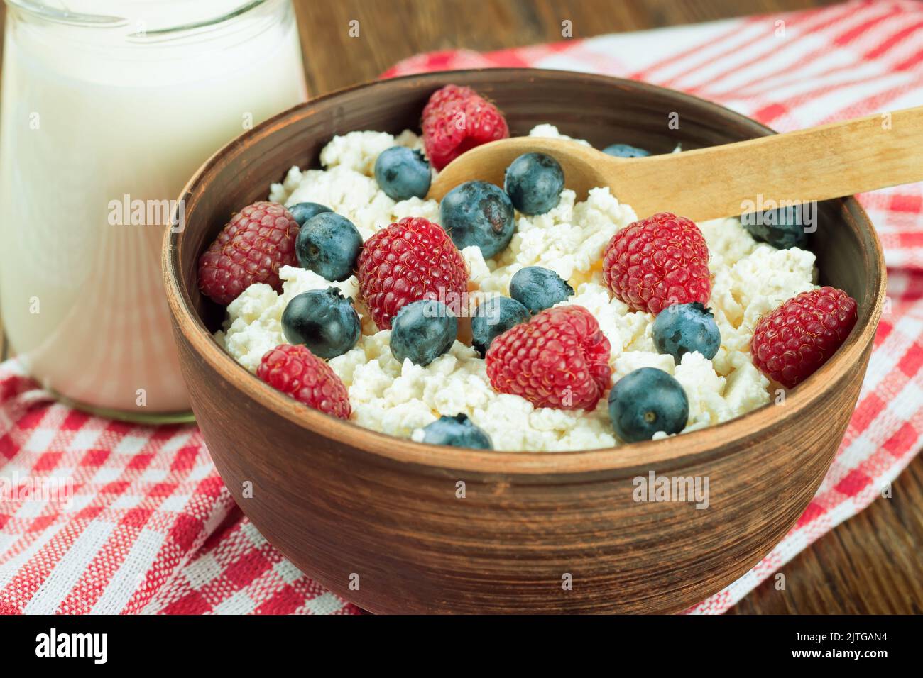 Quark mit Himbeeren und Heidelbeeren in brauner Tonschüssel und Holzlöffel auf rotem kariertem Handtuch. Milchprodukte, gesunde Lebensmittel. Stockfoto