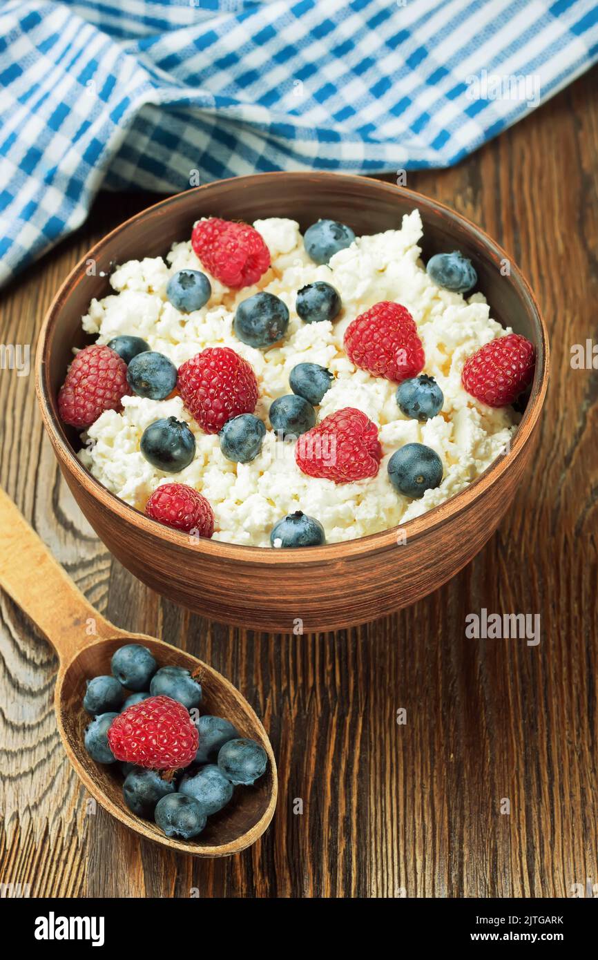 Quark mit Himbeeren und Heidelbeeren in brauner Tonschüssel und Holzlöffel auf braunem Holzhintergrund. Milchprodukte, gesunde Lebensmittel. Stockfoto