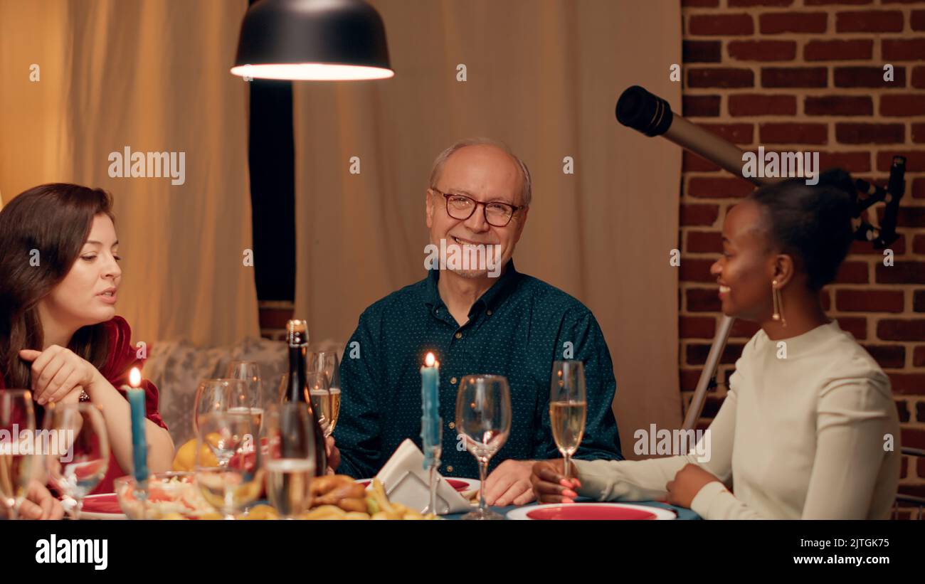 Fröhlicher älterer Mann, der sich mit einer jungen Frau unterhielt, während er am Weihnachtstisch mit Angehörigen zusammen war. Fröhlicher älterer Mensch, der mit seiner Tochter spricht, während er den Winterurlaub zu Hause gemeinsam feiert. Handgeführte Aufnahme Stockfoto