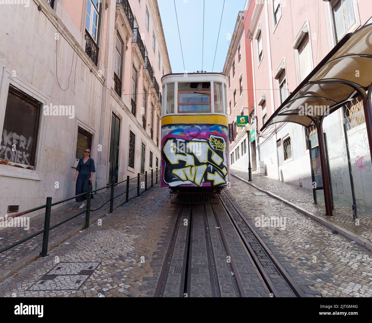 Steile Lissabonner Straße mit Straßenbahn alias Straßenbahn alias Trolley neben der Straßenbahnhaltestelle, während eine Frau die Straße entlang läuft. Sommer. Portugal Stockfoto