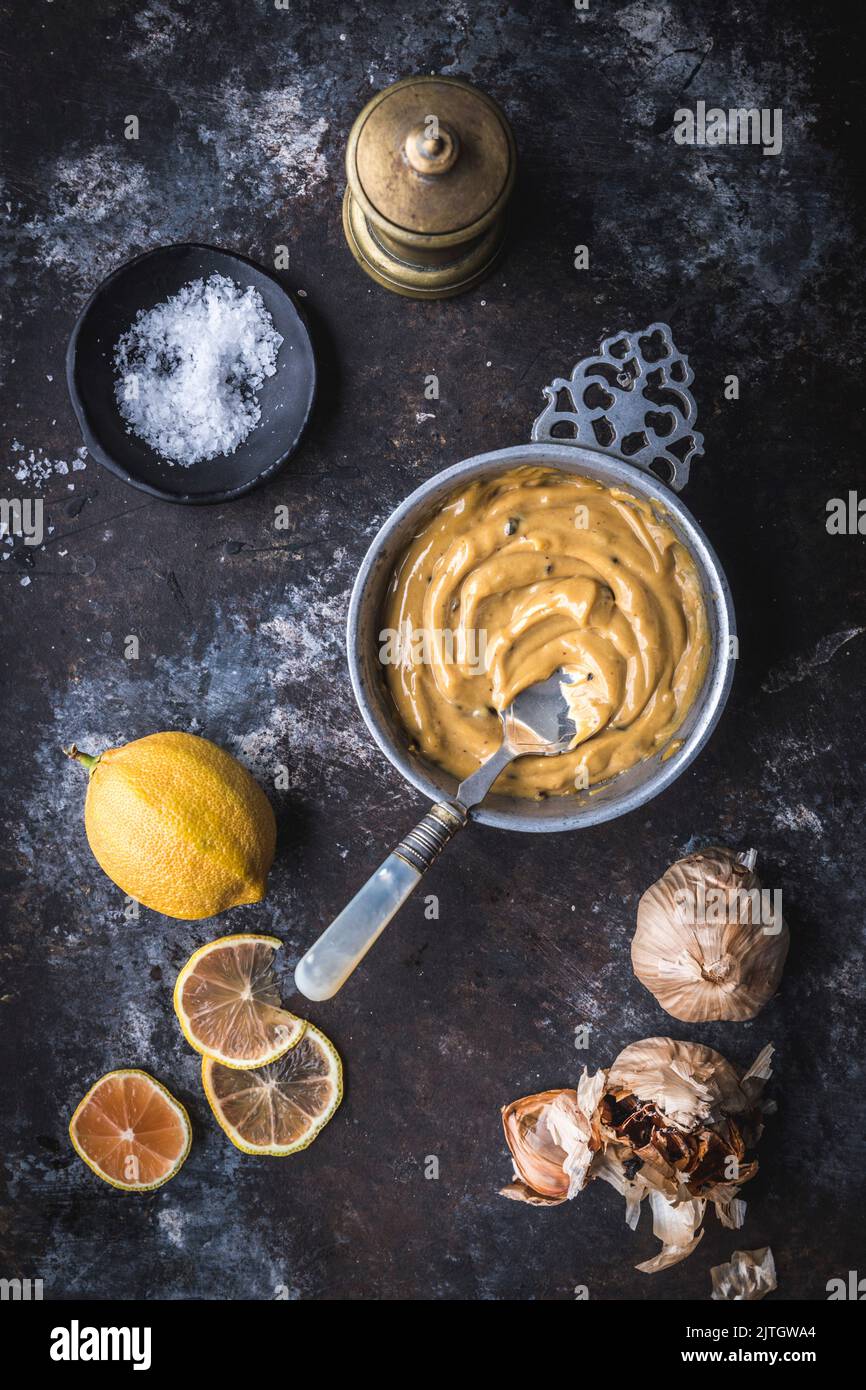 Knoblauch und Trüffel-Aioli in einer Metallschüssel mit rosa Zitronen, Knoblauch, Meersalz und Pfeffermühle Stockfoto