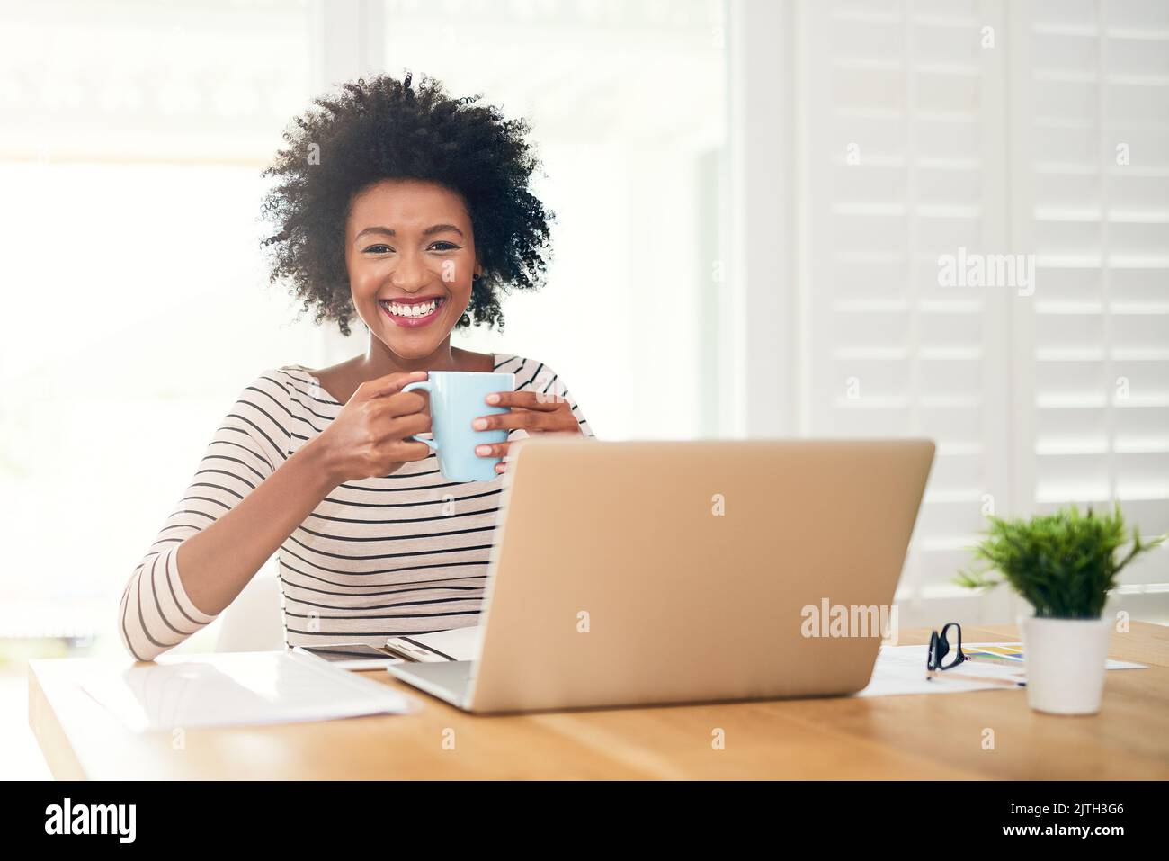 Ich kann nicht loslegen, ohne vorher eine gute Tasse Kaffee zu haben. Porträt einer jungen Frau, die Kaffee trinkt, während sie zu Hause an ihrem Laptop arbeitet. Stockfoto