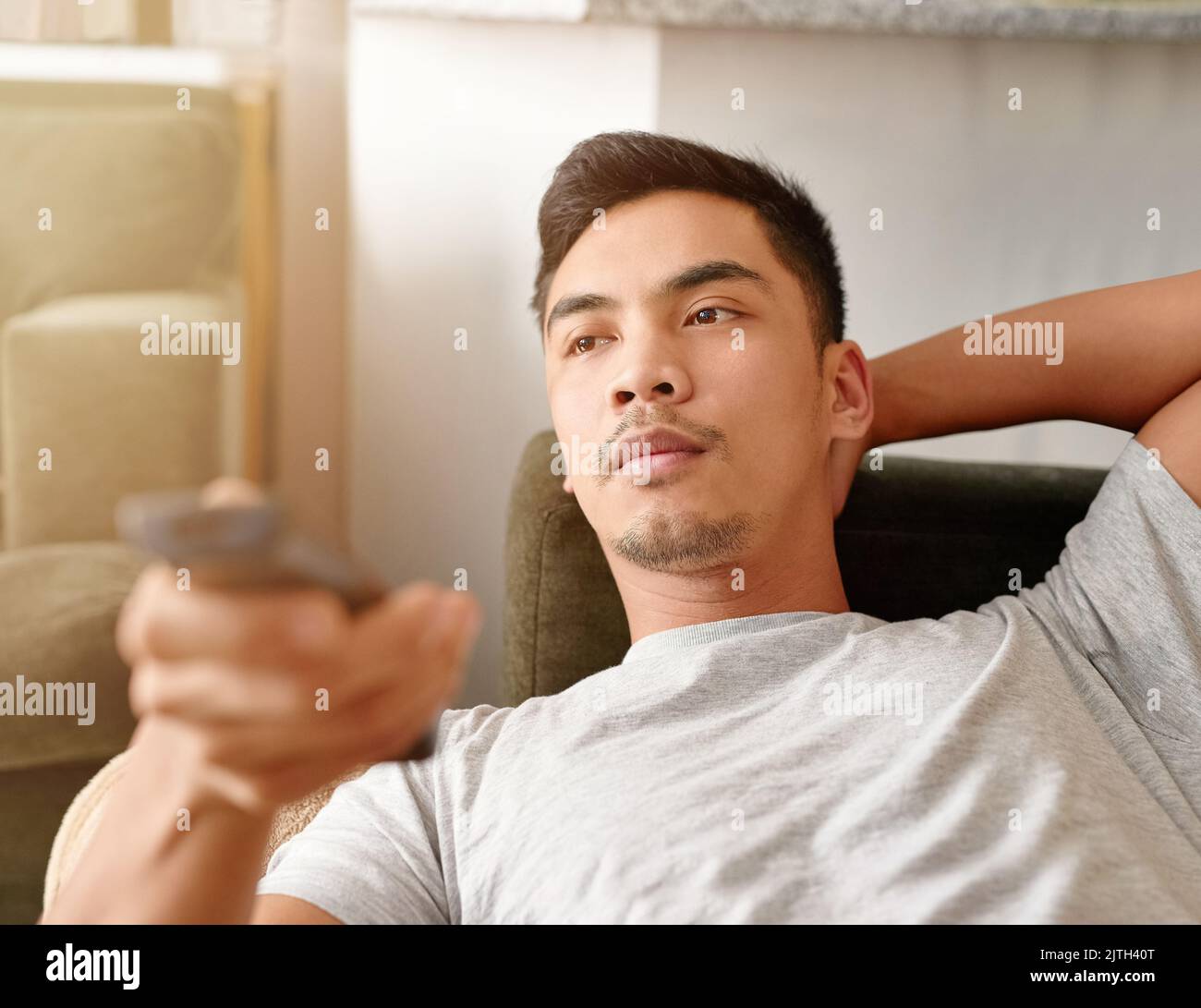 Samstags sind seine Couch Potato Tage. Ein junger Mann mit einer Fernbedienung, um den Kanal auf seinem Fernseher zu ändern, während er auf der Couch liegt. Stockfoto