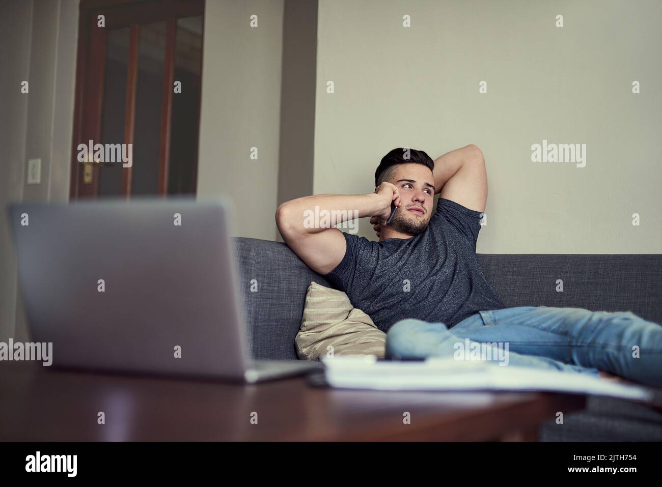 Zeit für eine kurze Studienpause. Ein fahrender junger Mann nimmt sein Telefon an, während er von seiner Lounge zu Hause aus arbeitet. Stockfoto