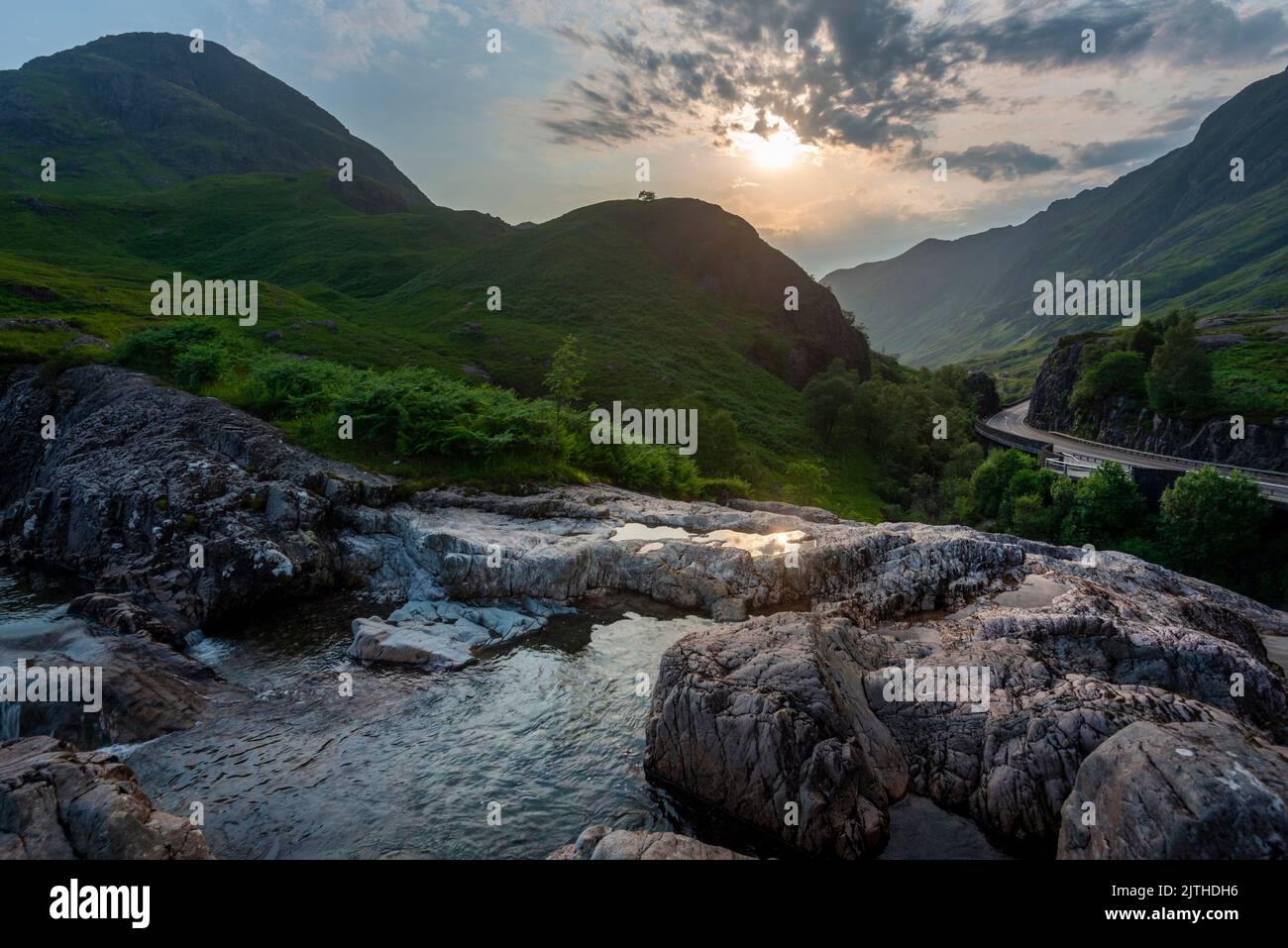 Die untergehende Sonne, über dem dramatischen Tal von Glen Coe, im Sommer, wo sich drei kleine Flüsse treffen, am Fuße der Three Sisters of Glen Coe, mit AW Stockfoto
