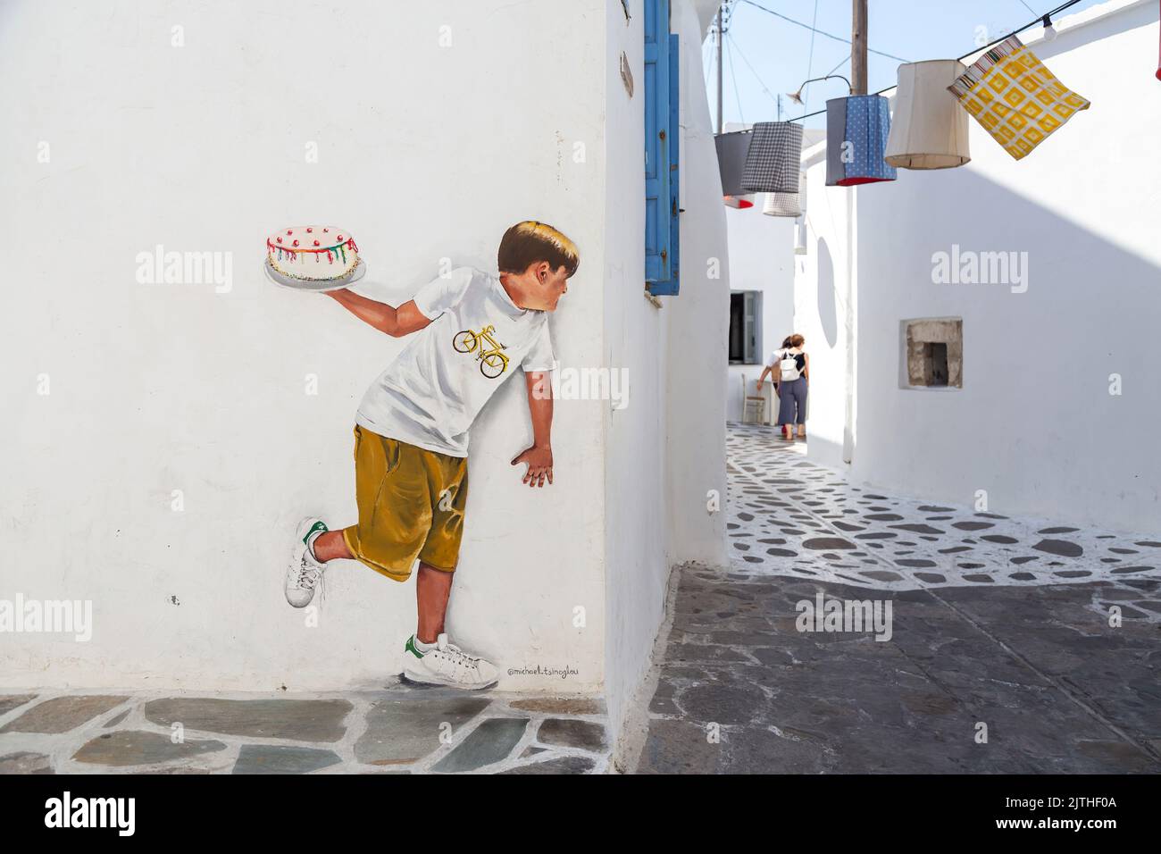 26. Juli 2022. Leben Sie wie ein Kind virale Graffiti an weiß getünchten kykladischen Wand. Das Kind hält einen Kuchen, um ihn dem Passanten zu werfen. Street Art Stockfoto