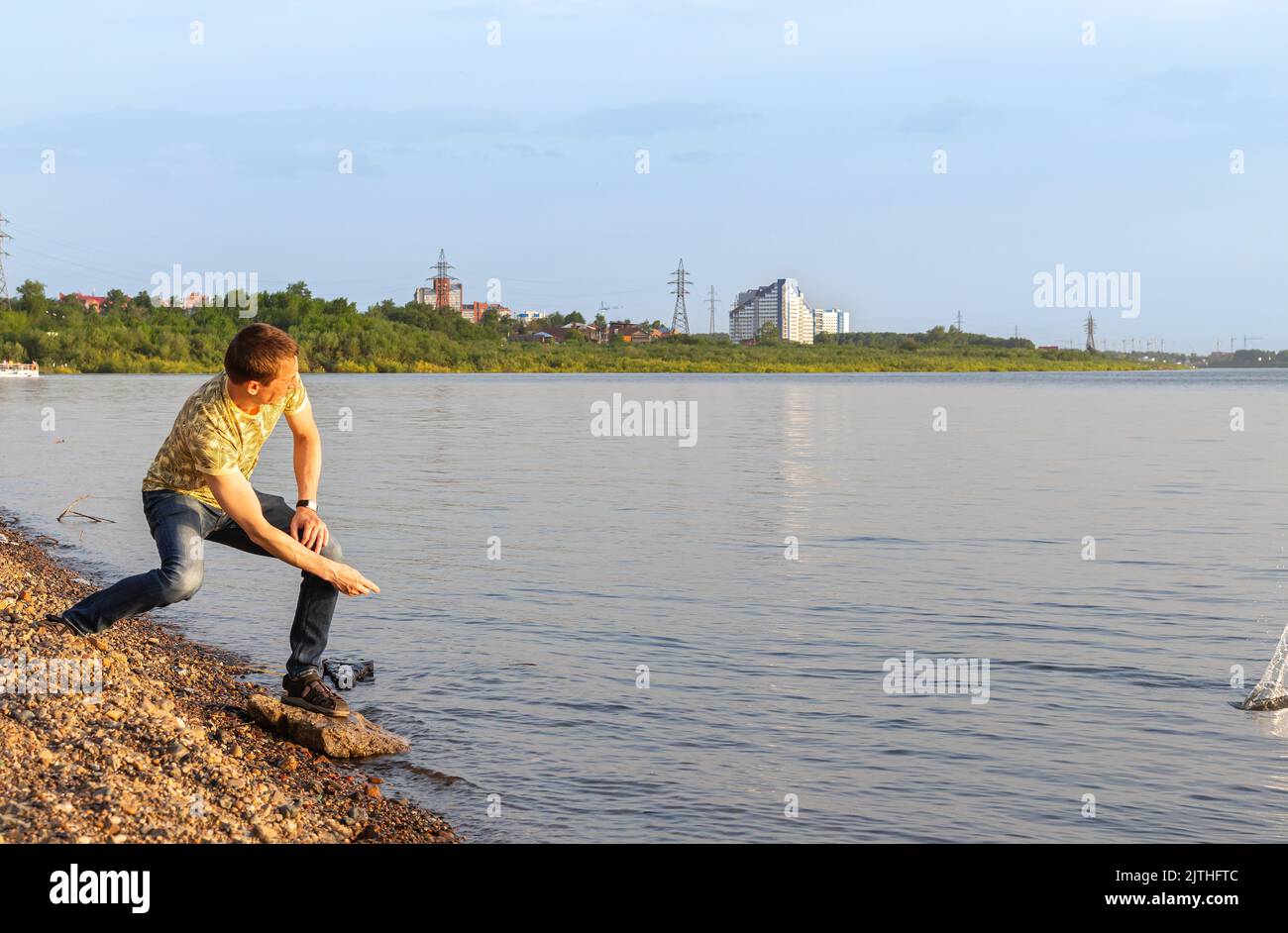 Junger Mann in gelbem T-Shirt und Jeans am Steinstrand, beim Steinspringen am Fluss. Schöner Mann in Sonnenstrahlen an warmen klaren Sommerabend Skipping Stones Stockfoto