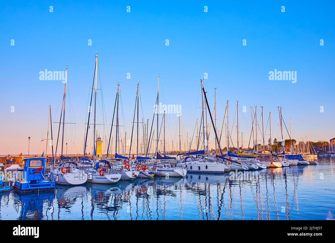 Die Linie der vertäuten Segelyachten im Hafen mit einem Leuchtturm und Sonnenuntergang Himmel im Hintergrund, Desenzano del Garda, Gardasee, Italien Stockfoto