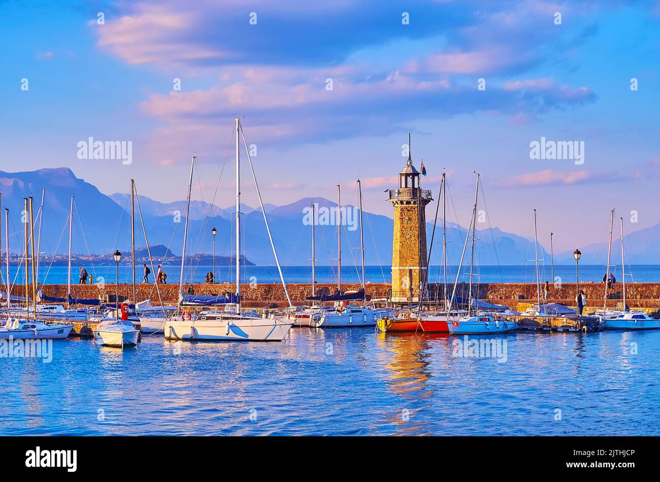 Genießen Sie den Spaziergang am Ufer des Gardasees mit Blick auf Yachten, Leuchtturm Faro, Garda Prealps, Desenzano del Garda, Lombardei, Italien Stockfoto