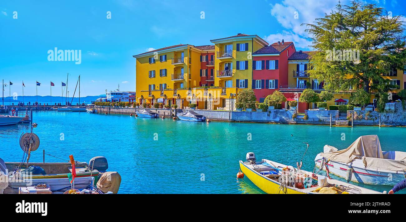 Panorama von kleinen farbigen Häusern und azurblauem Wasser von Sirmione Marina mit kleinen Booten, Fahnen und Linie von Garda Prealps im Hintergrund, Sirmione, Es Stockfoto