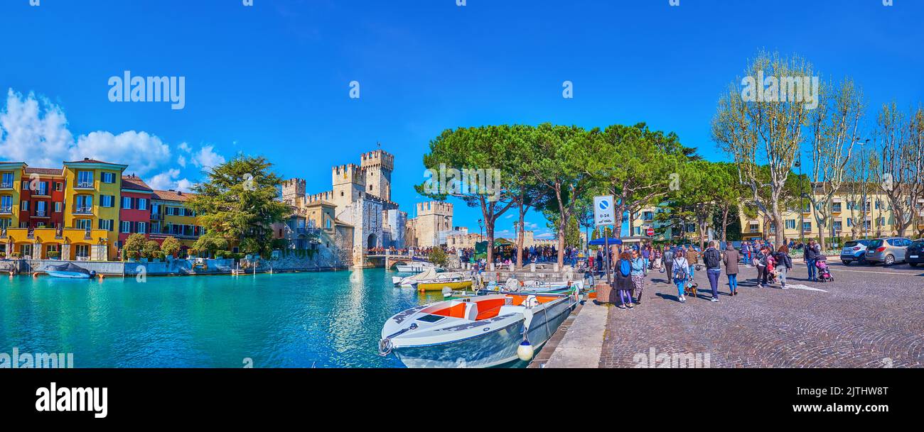 SIRMIONE, ITALIEN - 10. APRIL 2022: Panorama des malerischen Jachthafens mit festfahrenden Booten, Scaligero Castle, Sirmione Bridge, historischen Häusern und einem Blick auf L Stockfoto