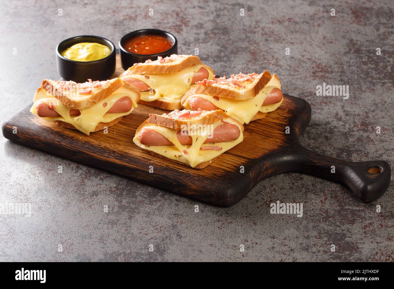 Hausgemachtes geröstetes Sandwich mit Wiener Würstchen und geschmolzenem Käse, serviert mit Ketchup und Senf aus nächster Nähe auf einem Holzbrett auf dem Tisch. Horizontal Stockfoto