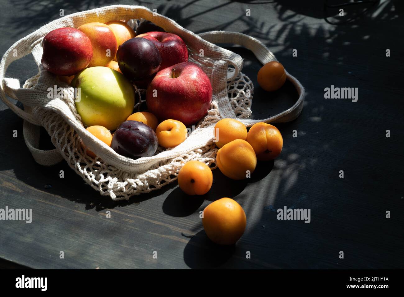 Frische reife Früchte und Beeren auf schwarzem Holzhintergrund in einem ökologischen Netzbeutel. Lifestyle. Harte Schatten.Sonnenlicht.reife Apfelernte. Kirschen, Aprikosen und Pflaumen. Speicherplatz kopieren. Stockfoto