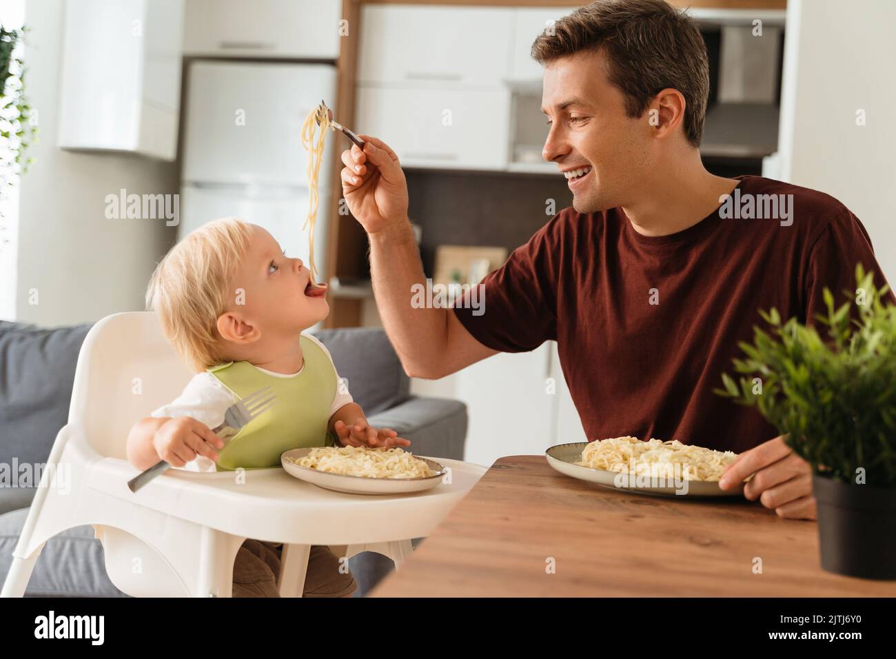 Alles gute zum Vatertag. Seitenansicht des Vaters, der das Baby in grünem Lätzchen mit Spaghetti während der Mittagszeit am Küchentisch füttert, das Kind sitzt auf einem Hochstuhl und hält die Gabel. Glückliche Vaterschaft, erste Mahlzeit, Selbsternährung Stockfoto
