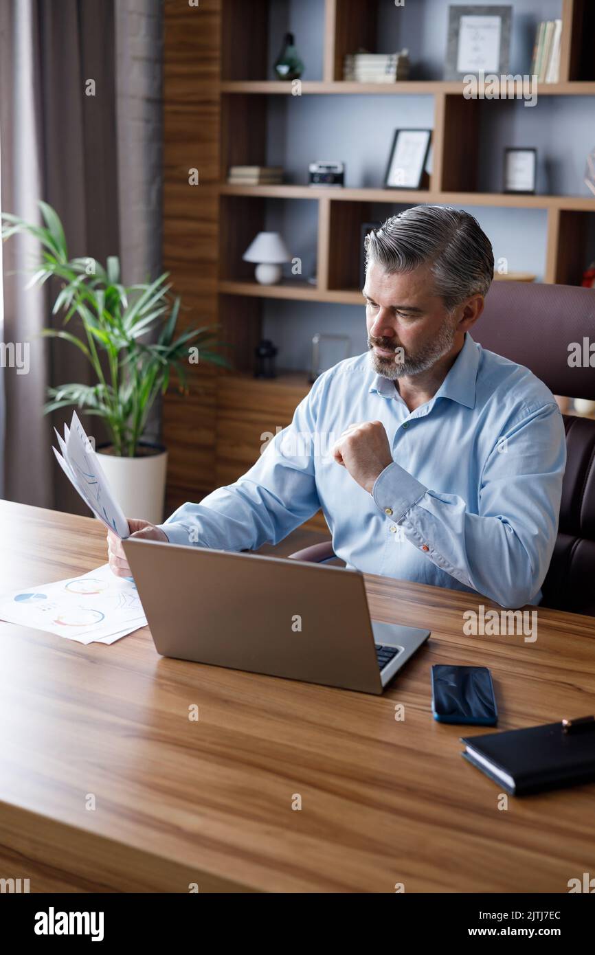 Porträt eines erwachsenen grauhaarigen bärtigen Geschäftsmannes, der im Heimbüro mit einem Laptop arbeitet. SMS senden, Schreibarbeiten erledigen, monatliche Ausgaben berechnen Stockfoto