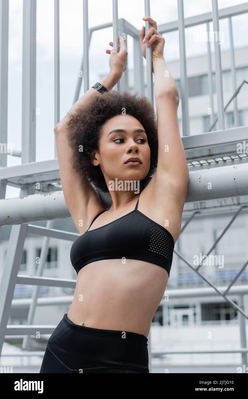 Hübsche afroamerikanische Frau mit schlankem Körper, die mit erhobenen Händen in der Nähe des Zauns posiert Stockfoto
