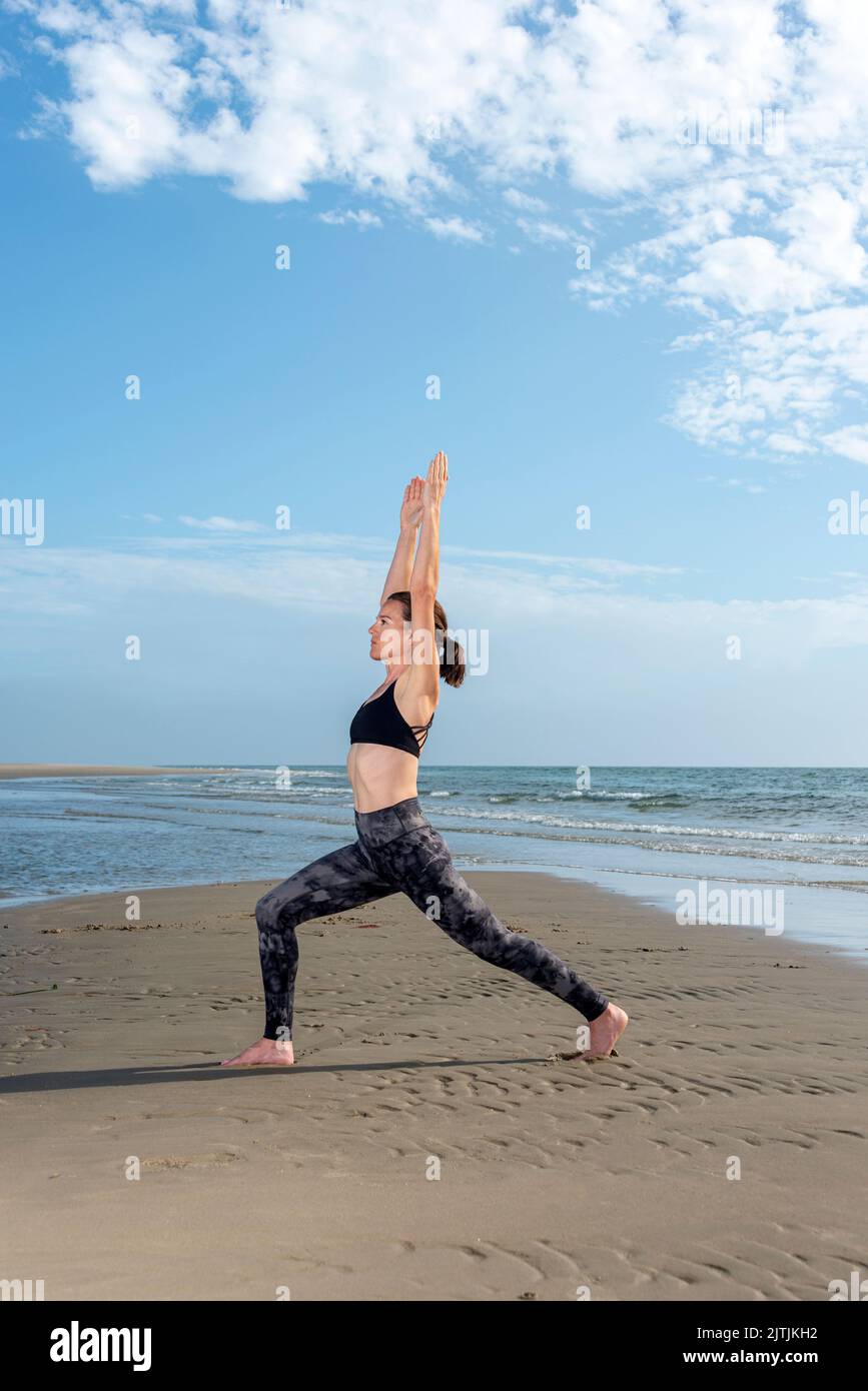 Sportliche Frau, die einen hohen Ausfallschritt beim Yoga macht, posiert am Strand in der Sonne Stockfoto