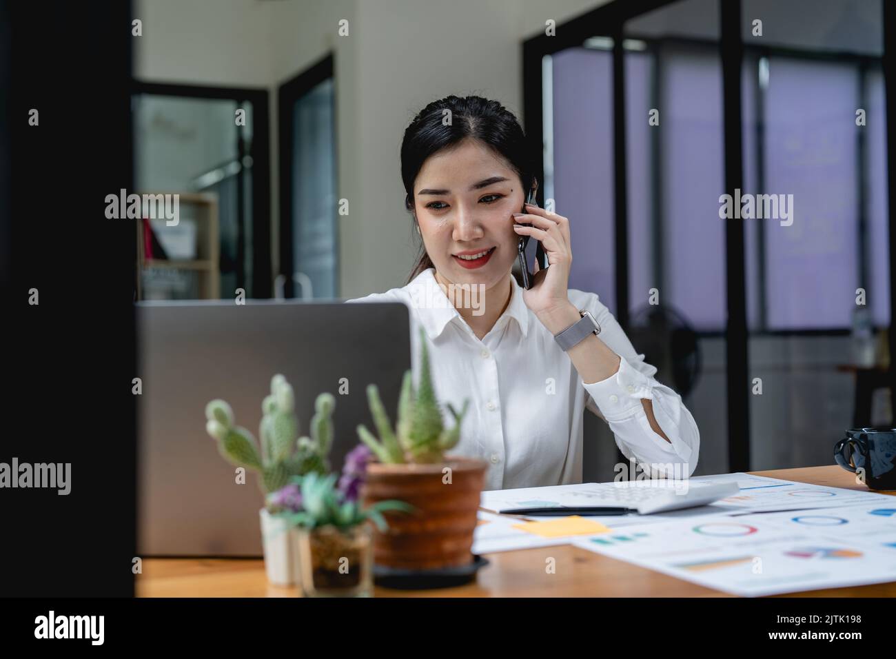 Junge asiatische Frau, die am Mobiltelefon spricht und lächelt, während sie an ihrem Arbeitsplatz im Büro sitzt. Stockfoto