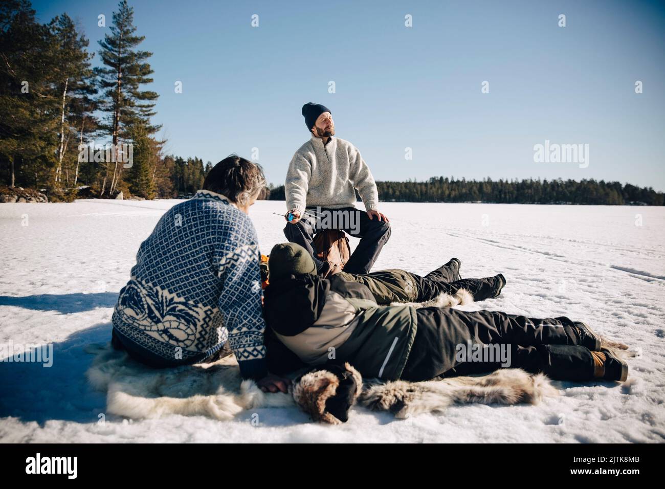 Vater und Sohn sitzen auf Tierhaut, während sie mit Freunden Eisfischen und im Winter sonnigen Tag genießen Stockfoto