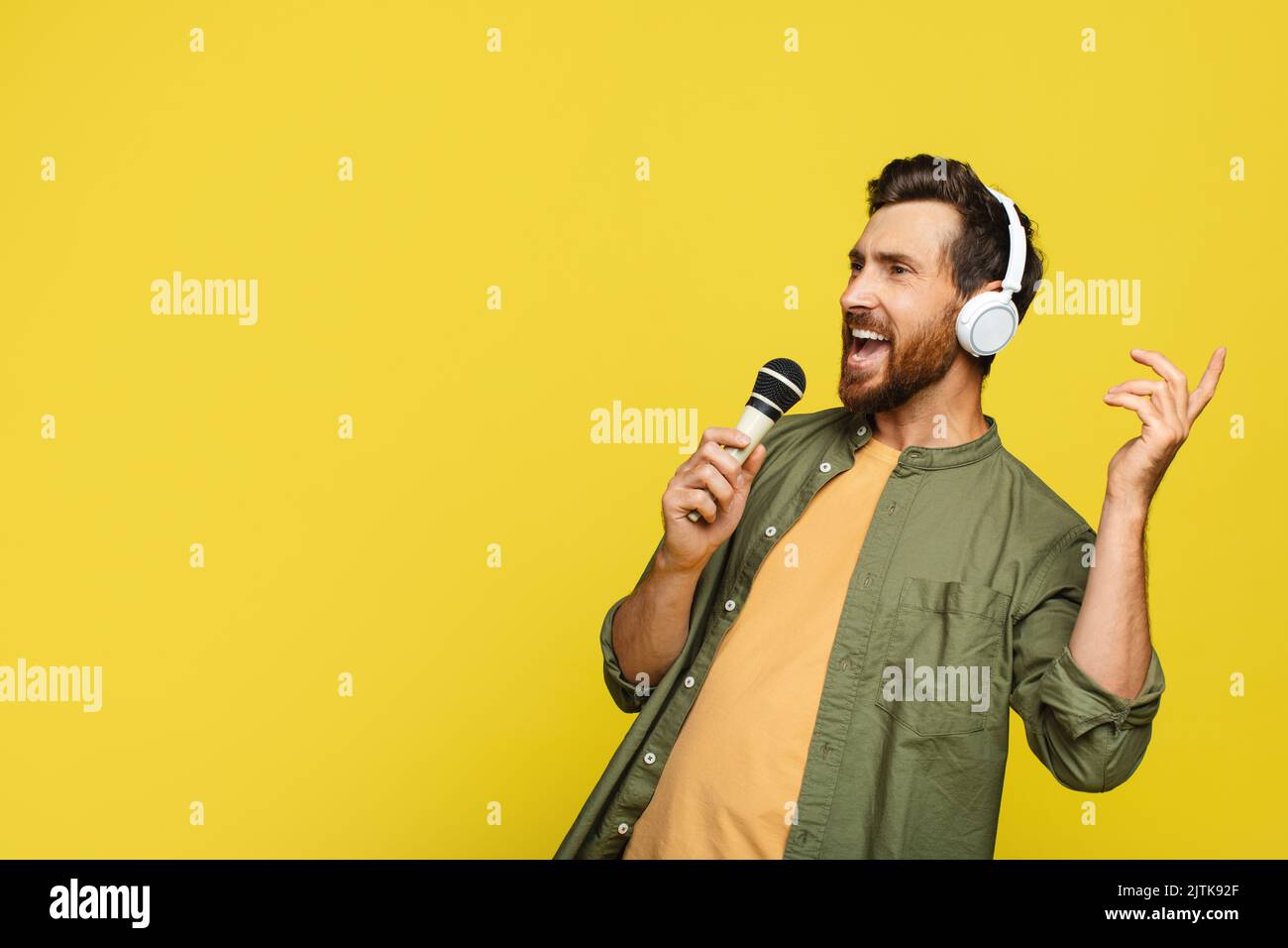 Fröhlicher Mann mittleren Alters in Kopfhörern, der Musik hört und Lied ins Mikrofon singt, gelber Hintergrund, Kopierraum Stockfoto