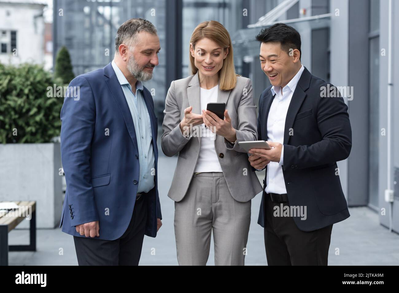 Drei UNTERNEHMER AUS DEM IT-Bereich vor dem Bürogebäude sehen sich am Telefon eines Kollegen ein Video an, glücklich und lächelnd, während sie sich in der Pause von der Arbeit ausruhen Stockfoto