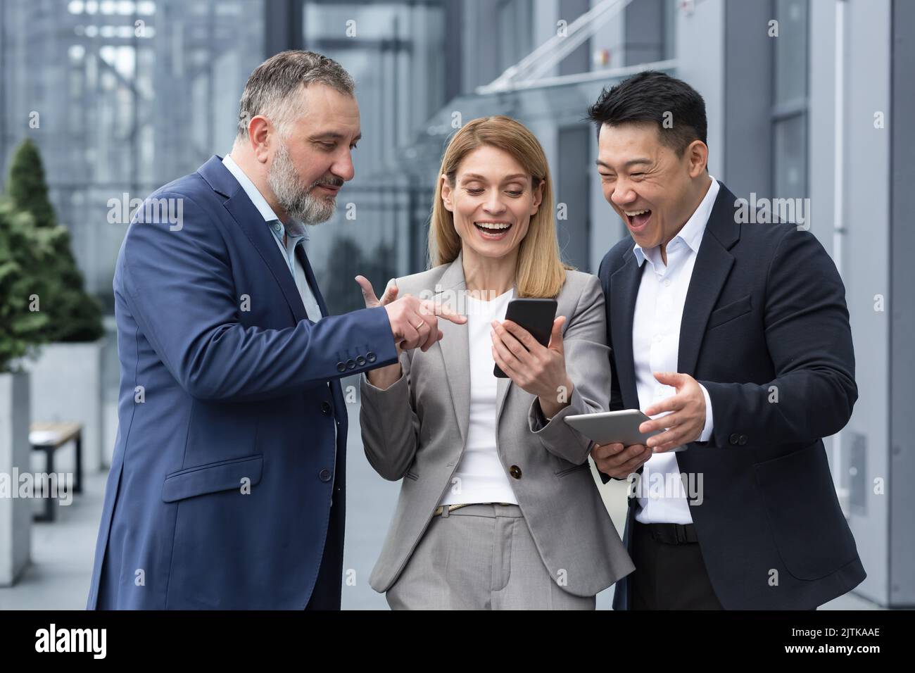 Drei UNTERNEHMER AUS DEM IT-Bereich vor dem Bürogebäude sehen sich am Telefon eines Kollegen ein Video an, glücklich und lächelnd, während sie sich in der Pause von der Arbeit ausruhen Stockfoto
