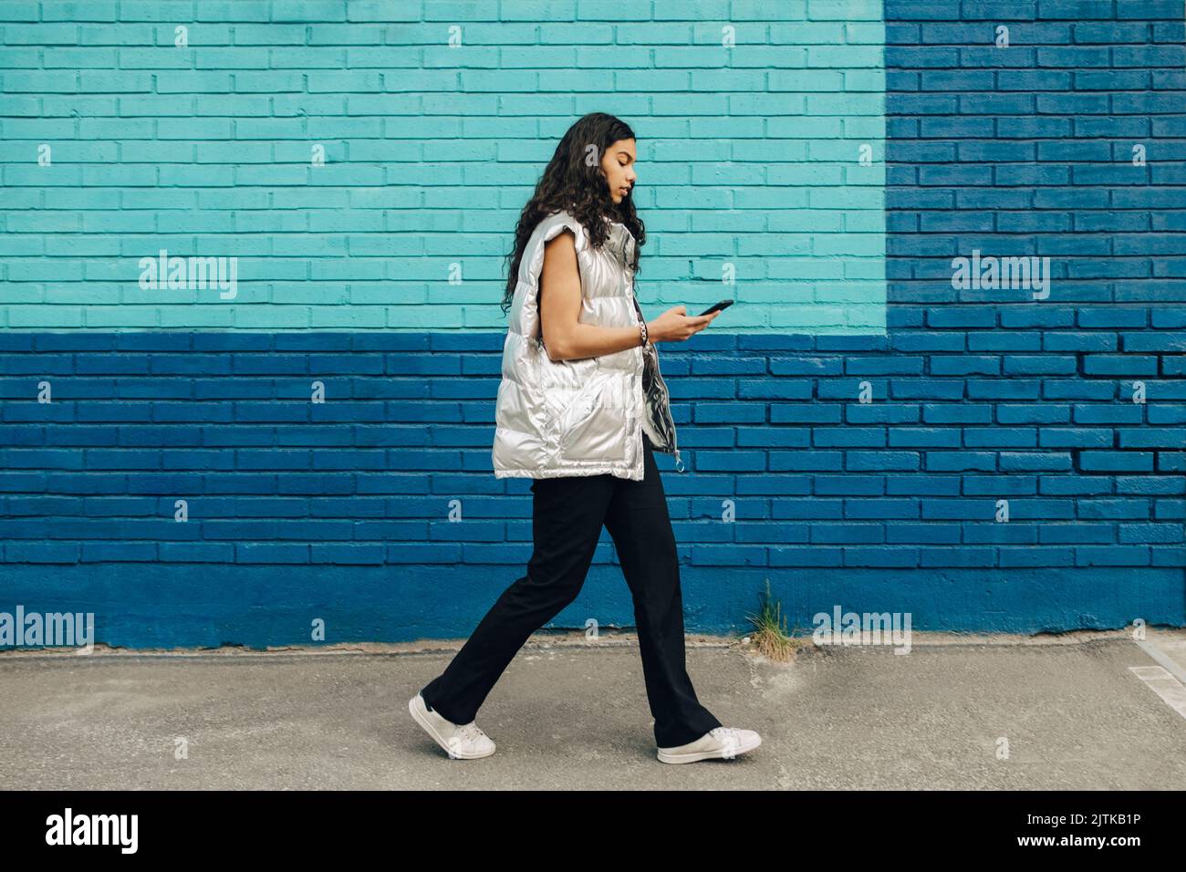 Junge Frau, die ein Mobiltelefon benutzt, läuft an einer blauen Ziegelwand vorbei Stockfoto