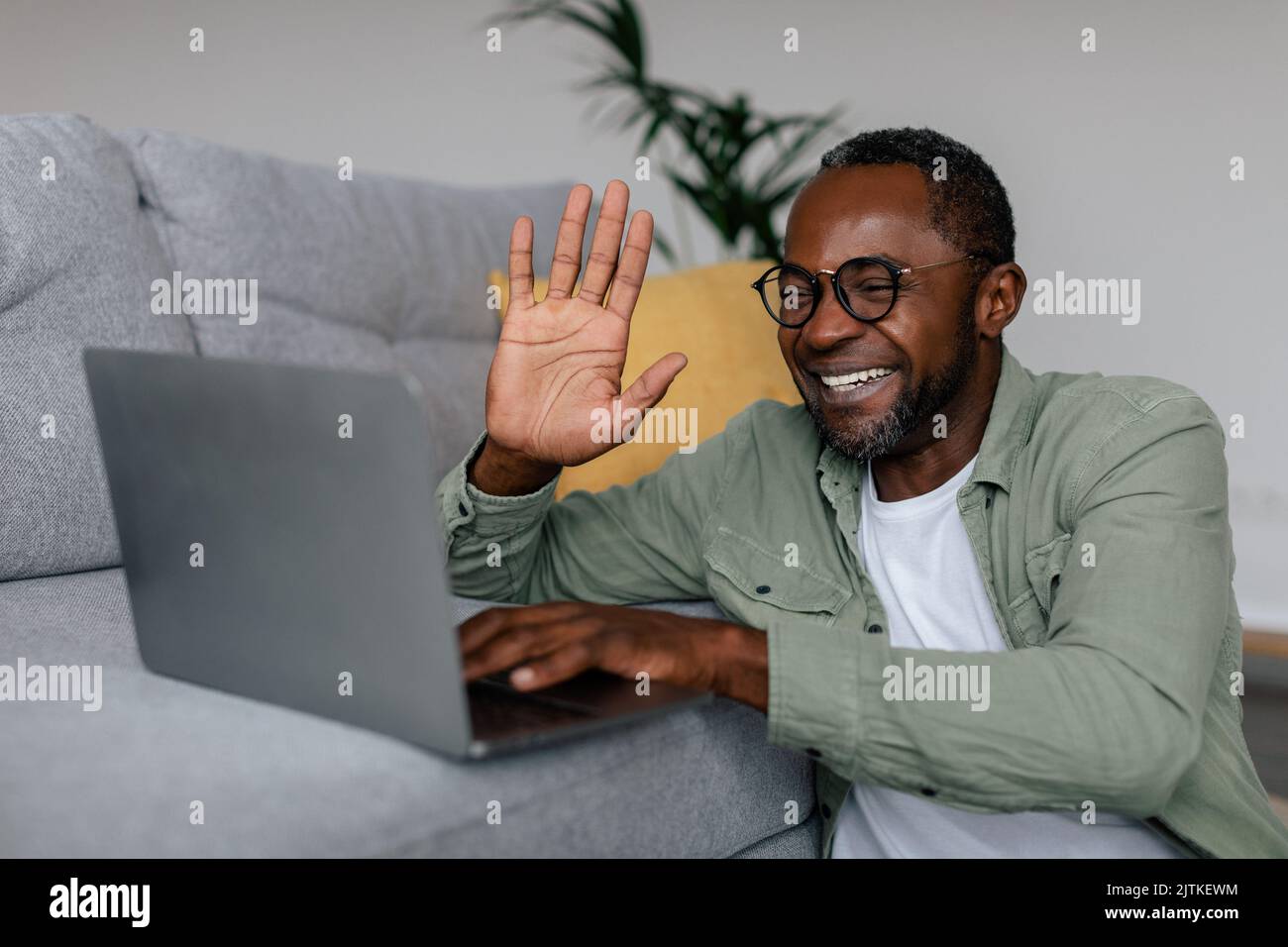 Lächelnder afroamerikanischer Mann mit Brille, der mit der Hand auf den Laptop im Wohnzimmer schwenkt Stockfoto