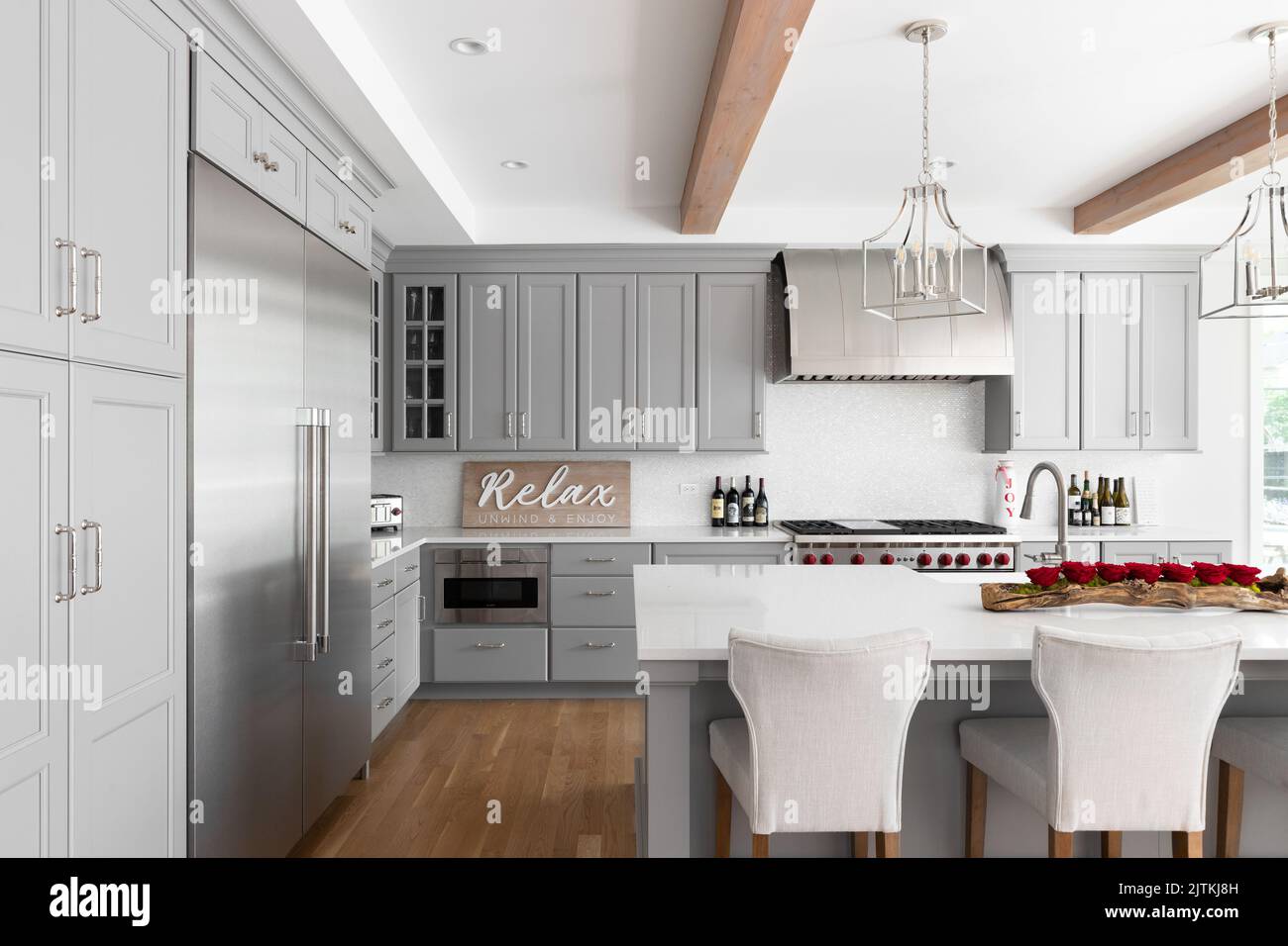 Eine luxuriöse Küche mit grauen Schränken und großer Insel, Arbeitsflächen aus weißem Marmor, freiliegenden Holzbalken und Edelstahlgeräten. Stockfoto