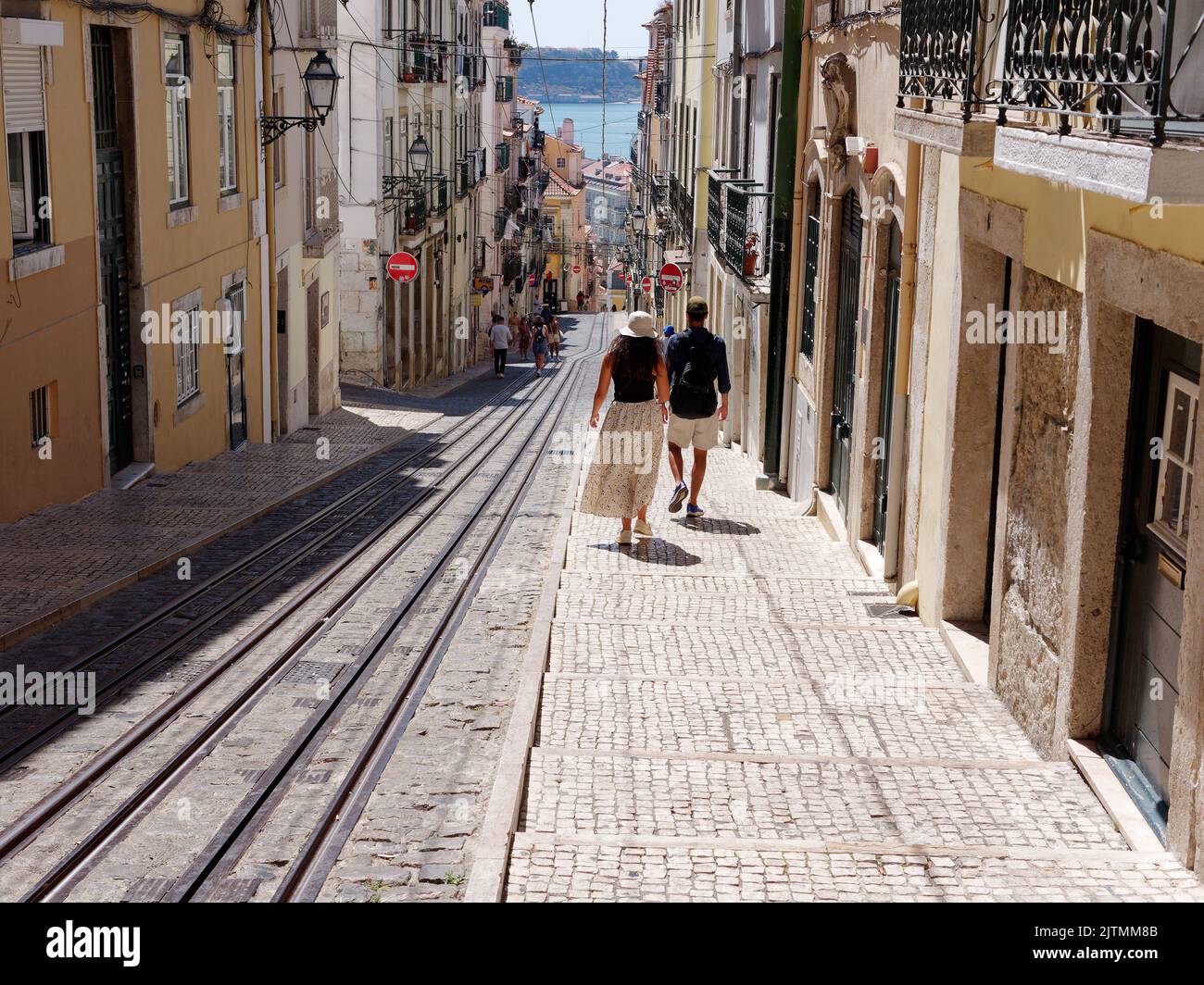 Ein paar gehen an einem Sommertag mit dem Fluss Tejo in der Ferne eine steile Straße mit Straßenbahnlinien in der Gegend Baixa-Chiado von Lissabon hinunter. Portugal. Stockfoto