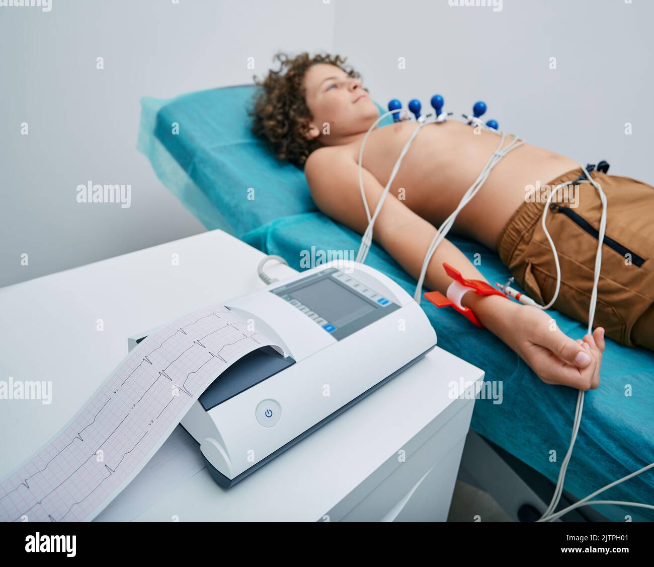 EKG-Schreiber mit EKG-Ausdruck für einen Teenager-Jungen, der im medizinischen Bett mit Vakuumsensoren liegt. Herzelektrokardiographie für Kinder Stockfoto