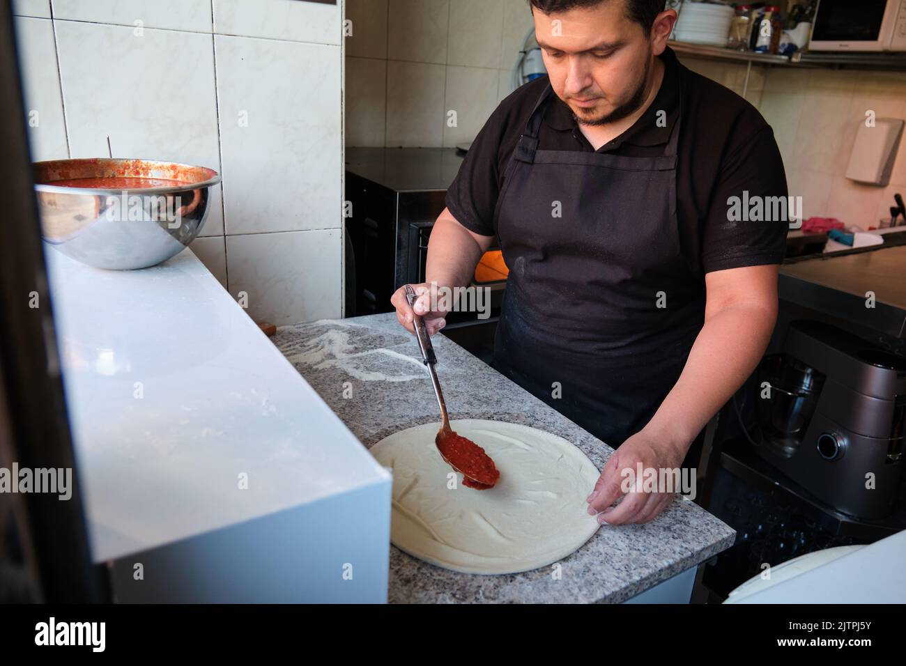 Lateinischer Mann, der in einer Restaurantküche Tomatensauce zu einem Pizzateig hinzufügt. Stockfoto