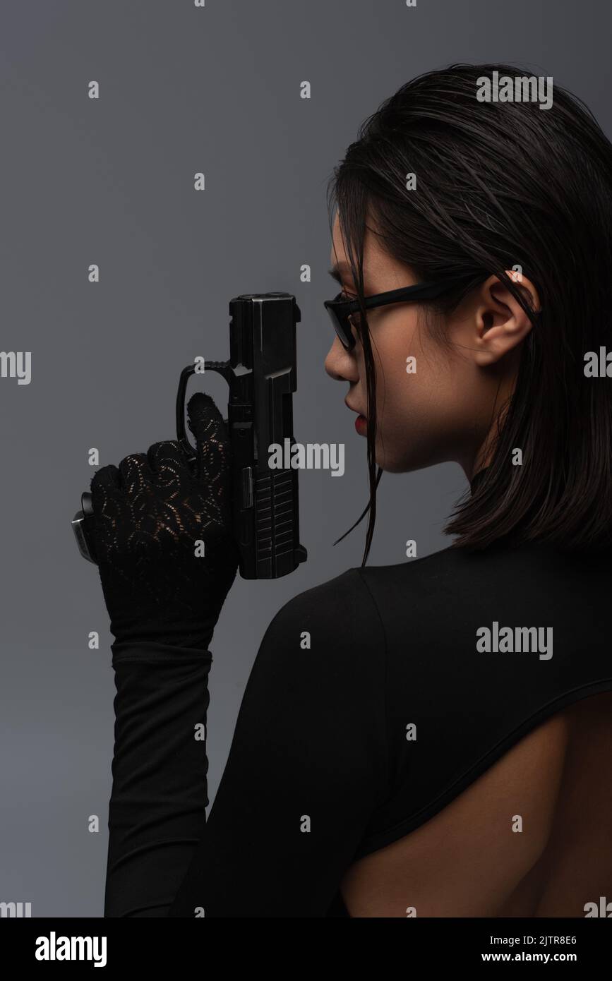 Seitenansicht der gefährlichen asiatischen Frau in total schwarzen Outfit und stilvolle Sonnenbrille halten Waffe isoliert auf grau, Stock Bild Stockfoto