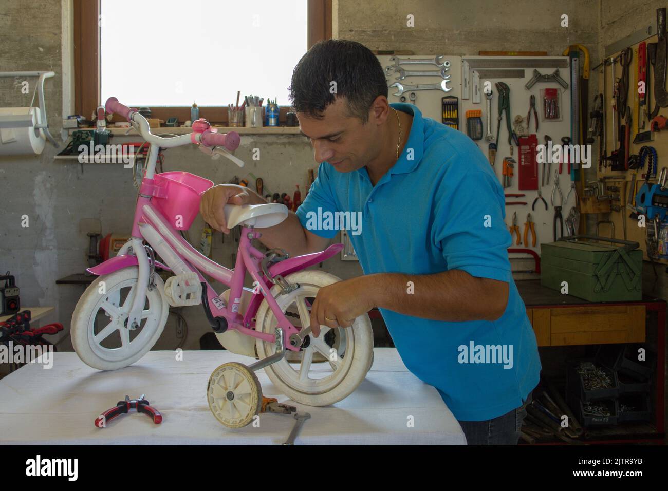 Bild eines jungen Handwerkerdades, der in seiner Werkstatt das Fahrrad seiner Tochter repariert. Do-it-yourself-Arbeit Stockfoto