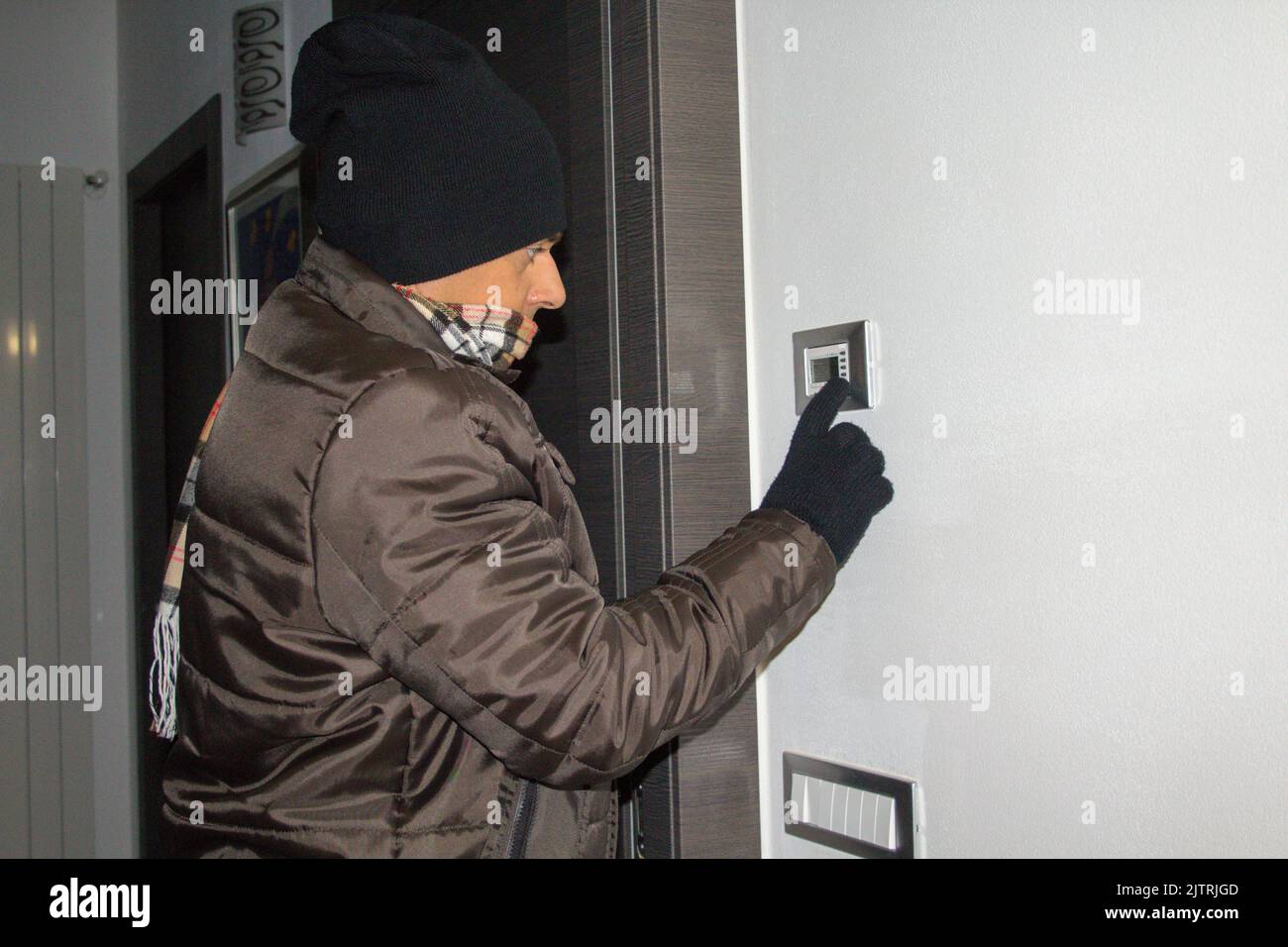 Mann in Jacke, Schal, Handschuhen und Wollmütze fühlt sich zu Hause kalt an, während er den Heizungsthermostat aufdreht. Hinweis auf den kommenden Winter Stockfoto