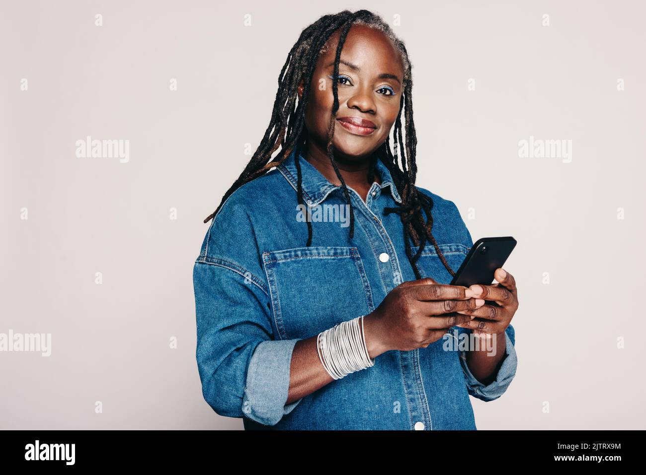 Schöne schwarze Frau, die auf die Kamera schaut, während sie ein Smartphone hält und vor einem grauen Hintergrund steht. Reife Frau mit Dreadlocks trägt ein Stockfoto