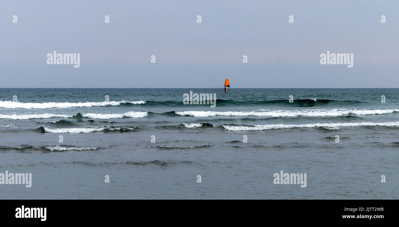 Grauer Himmel über dem Meer. seascape. Windsurfen. Ein Brett mit einem orangefarbenen Segel. Surfen auf dem kalten Meer. Person, die auf Meereswellen surft Stockfoto