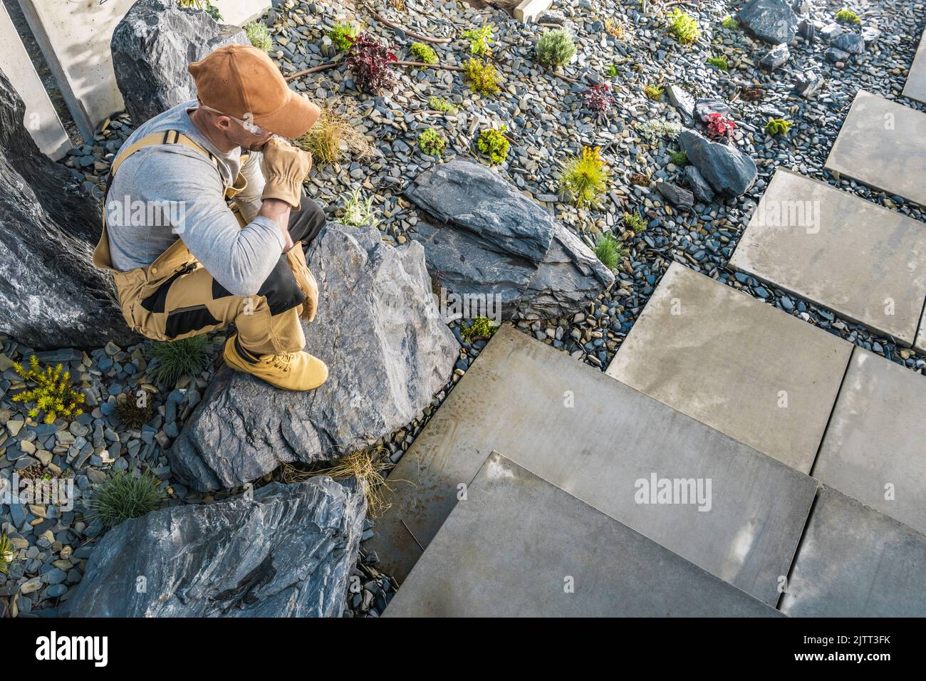 Landschaftsgärtner sitzt auf einem dekorativen Stein im Garten seines Kunden und beobachtet und analysiert die Gestaltung des Yards, um die Landschaftsbau- und Designarbeiten zu planen. Stockfoto