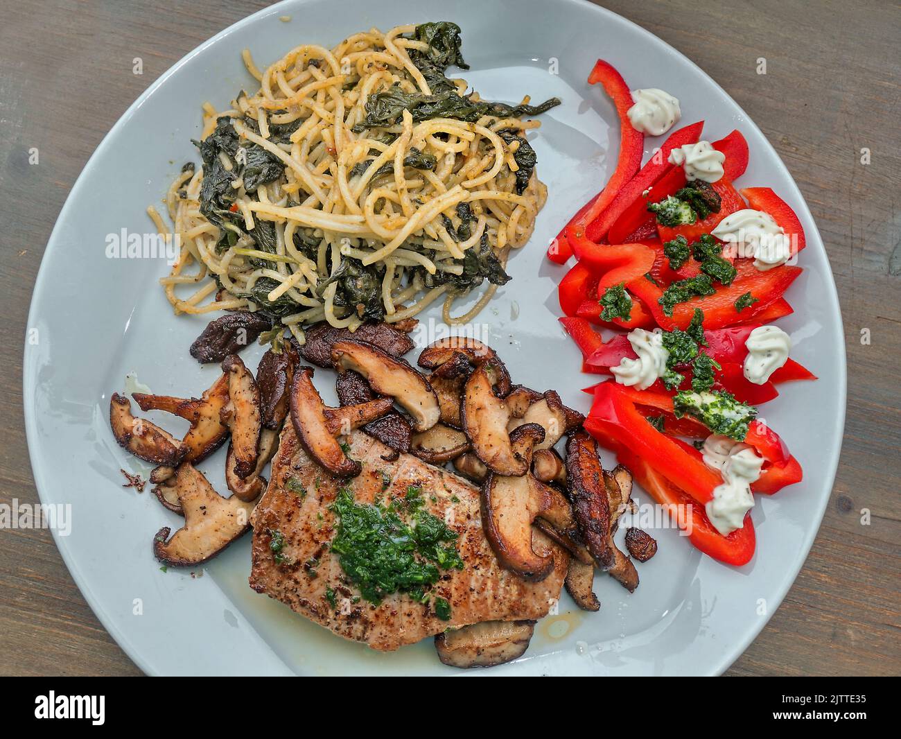 Draufsicht auf einen hellgrauen Essteller mit gebratenem Thunfisch, gerösteten Shiitake-Pilzen, Spaghetti mit Kapern und Spinatblättern und rohem roten Paprika Stockfoto