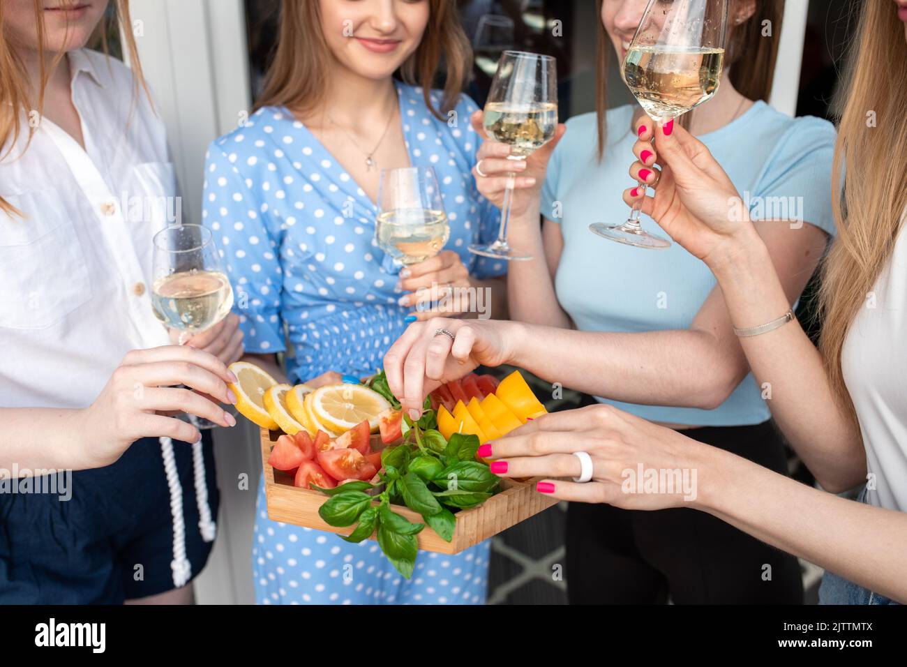Beschnittenes Foto von jungen lächelnden atemberaubenden Frauen, die Gemüse halten, in der Nähe des Hauses klirrende Gläser Weißwein halten. Stockfoto