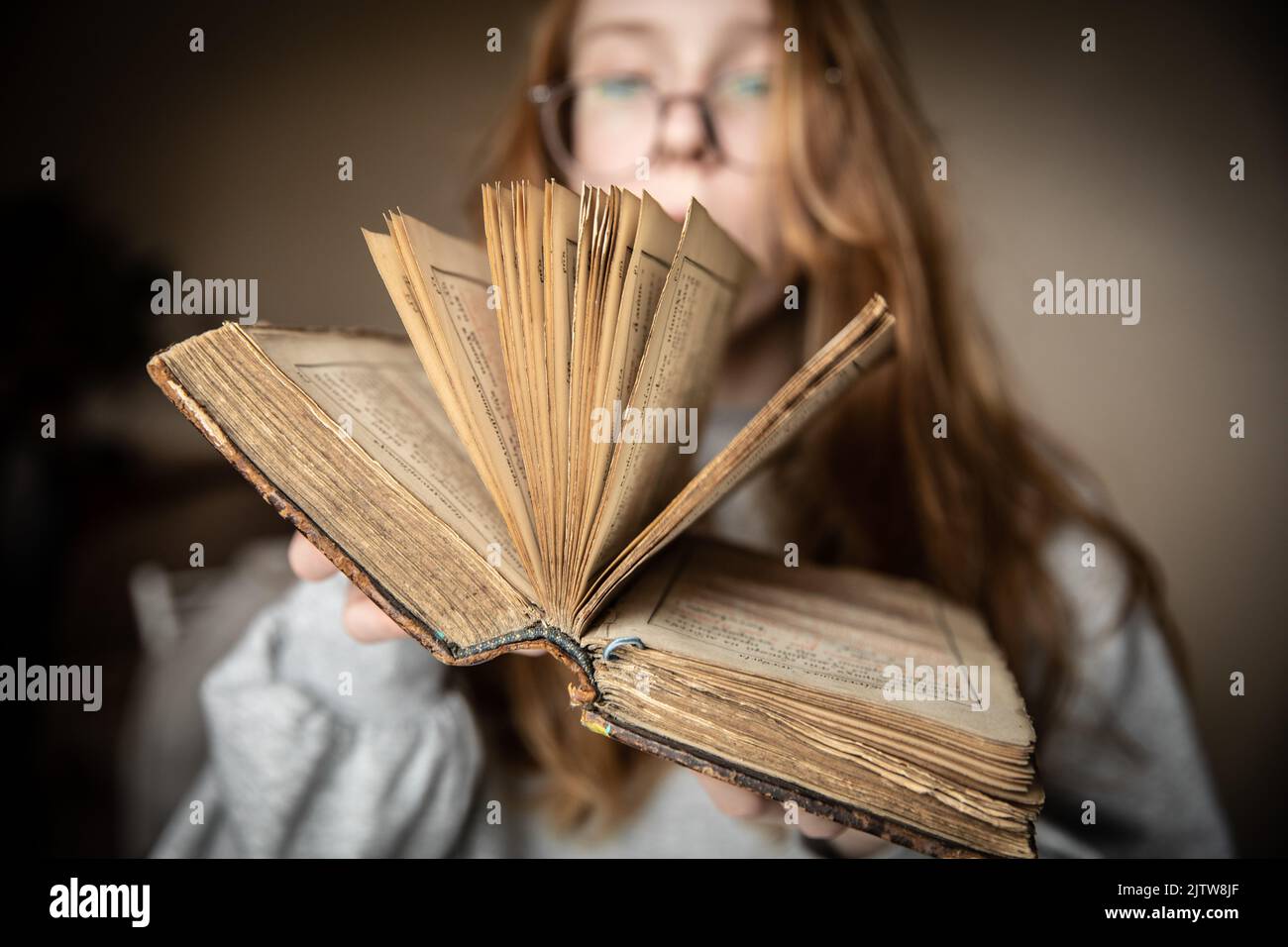 Ein offenes Vintage-Buch in den Händen eines niedlichen Teenagers mit Brille und roten Haaren in einem halbdunklen Raum, der vom Licht des Fensters beleuchtet wird Stockfoto