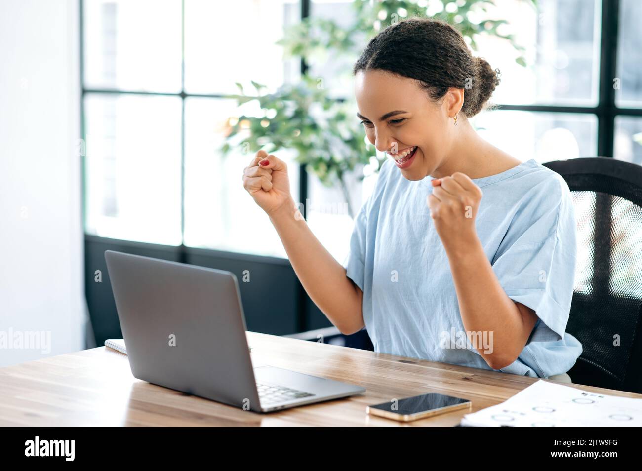 Freudig erstaunt glückliche brasilianische oder hispanische junge Geschäftsfrau, sitzt an einem Schreibtisch mit einem Laptop im modernen Büro, freut sich über Erfolg, großen Gewinn, feiern Deal, gestikulieren mit Fäusten, Lächelnd Stockfoto