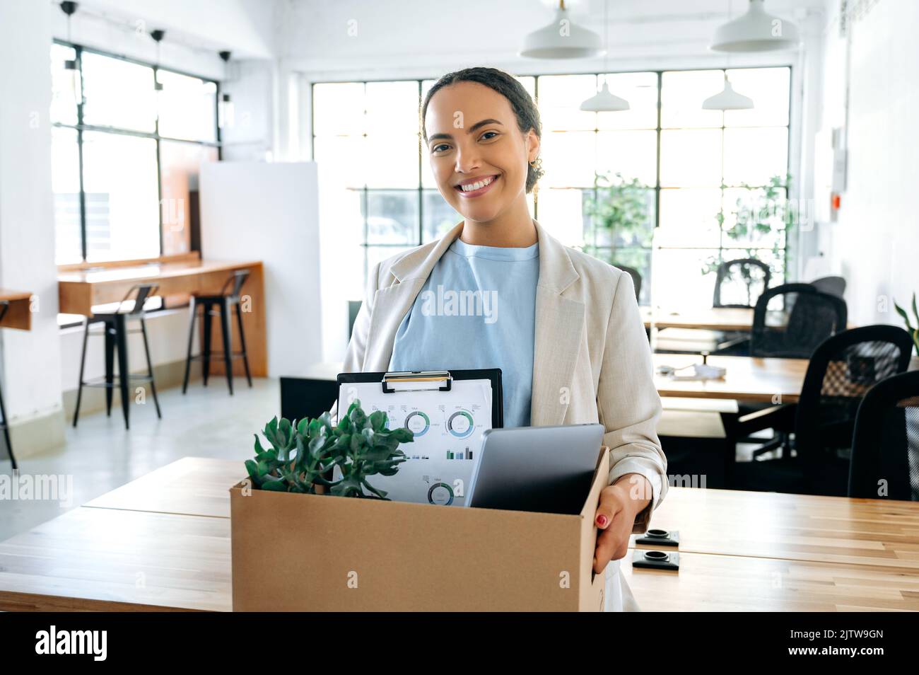 Glückliche junge brasilianische oder hispanische Mitarbeiterin, die einen Karton in der Hand hält, im modernen Büro steht und lächelnd auf die Kamera schaut, bekam einen neuen Job, ihren ersten Arbeitstag in einem neuen Arbeitsplatz Stockfoto