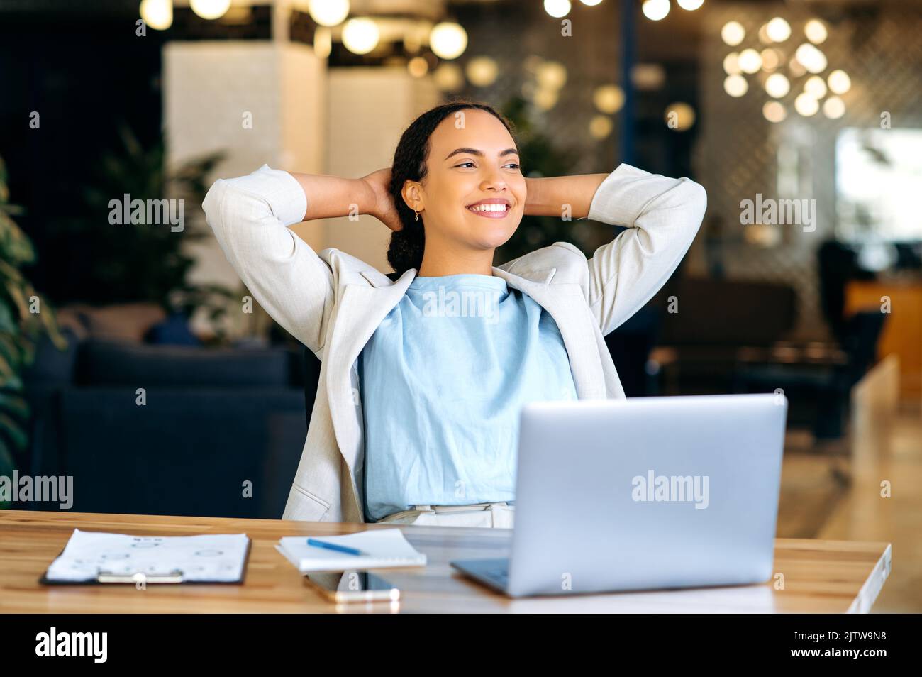 Ruhige, entspannte, gemischte Mitarbeiterin, die während der Arbeitszeit ausruht, im modernen Büro sitzt, die Hände hinter den Kopf stellt, sich mit dem Projekt zufrieden fühlt, die Arbeit befördert, in die Ferne blickt und freundlich lächelt Stockfoto