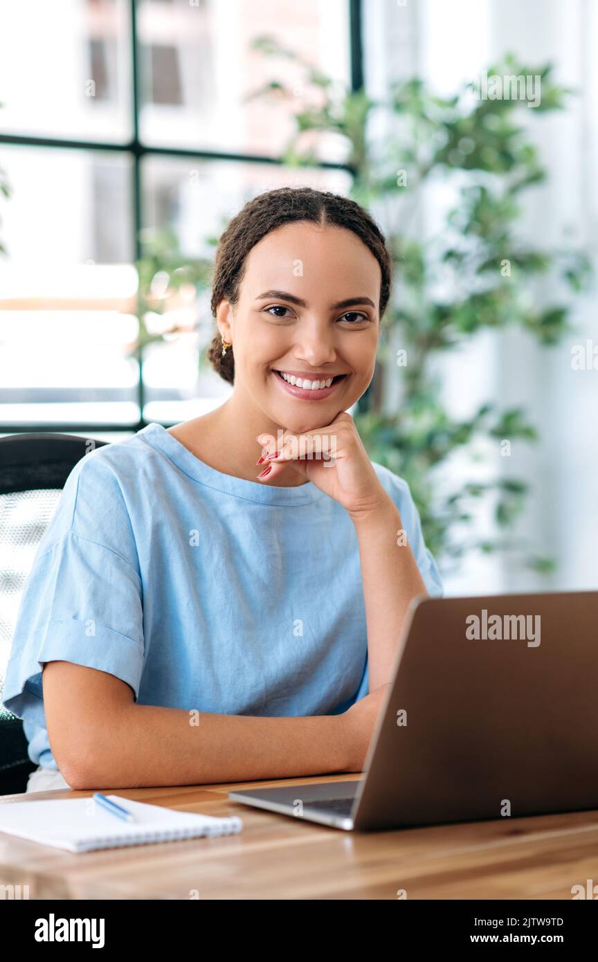 Vertikales Foto einer schönen, eleganten brasilianischen oder hispanischen jungen Frau, Maklerin, Kreativmanagerin, ceo-Firma, sitzt auf einem Schreibtisch mit Laptop in einem modernen Kreativbüro und schaut lächelnd auf die Kamera Stockfoto