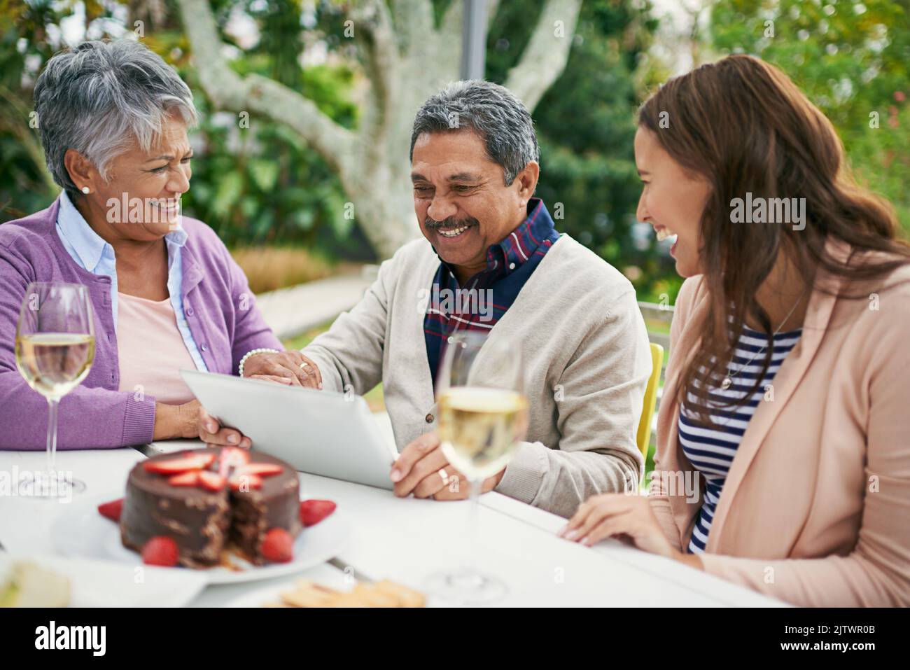 Aber wie schalte ich es ein. Ein älterer Mann zeigt seiner Familie etwas auf seinem Tablet, während sie draußen zu Mittag essen. Stockfoto